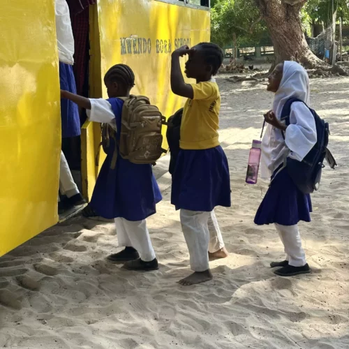 Drei Schulkinder mit Rucksäcken steigen an einer gelben Schulbus-Tür in Kenia ein, Sandboden und Bäume im Hintergrund.