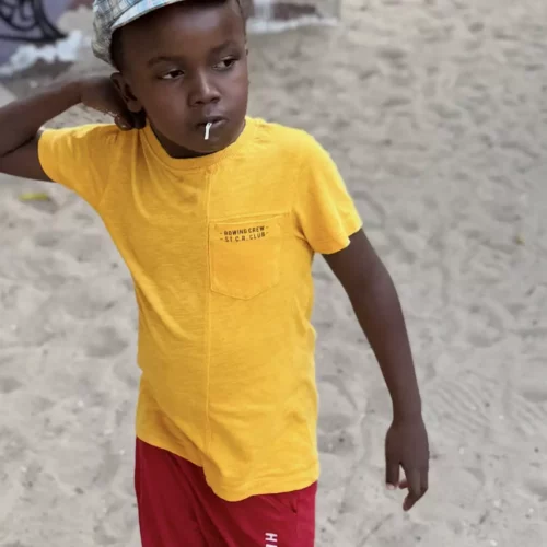Junge in gelbem T-Shirt, roter Shorts und Kappe steht nachdenklich im Sand vor bunter Wand mit Graffiti, Kenia.
